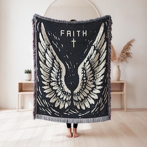 Puede incluir: Una manta tejida en blanco y negro que presenta un gran par de alas de ángel, la palabra FAITH y un pequeño símbolo de cruz centrado en la parte superior. La manta rectangular tiene flecos decorativos en los bordes superior e inferior.