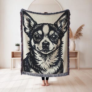 May include: A woven throw blanket featuring a high-contrast black and white graphic portrait of a Chihuahua dog. The rectangular blanket has a textured knit construction and is finished with decorative fringed edges along the top and bottom sides.