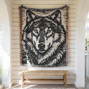 May include: A woven tapestry featuring a detailed black and white wolf portrait. The tapestry has a fringed edge with brown and tan accents. It is hanging on a white slatted wall above a wooden bench.