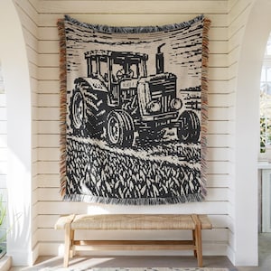Puede incluir: Una manta tejida con un diseño gráfico en blanco y negro de un tractor en un campo. El textil rectangular tiene un borde con flecos y hilos multicolores en los lados, mostrando un motivo detallado de un vehículo agrícola.
