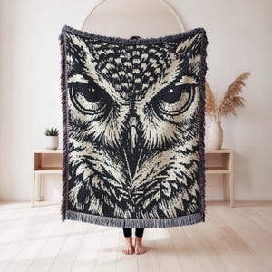 May include: A rectangular woven throw blanket featuring a high-contrast black and cream graphic design of an owl face. The blanket has a detailed, pixelated texture with a fringed edge along the top and bottom, showcasing a symmetrical pattern.