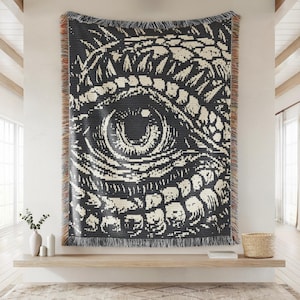 May include: Black and cream-colored woven throw blanket featuring a large eye design. The blanket has a fringed edge and is displayed on a wall. The eye design is detailed and graphic, creating a striking visual.