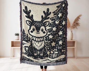 May include: A woven throw blanket featuring a black and cream graphic design of a cartoon reindeer with large antlers, standing next to a decorated Christmas tree with star ornaments and a gift box, finished with a fringed edge on all four sides.