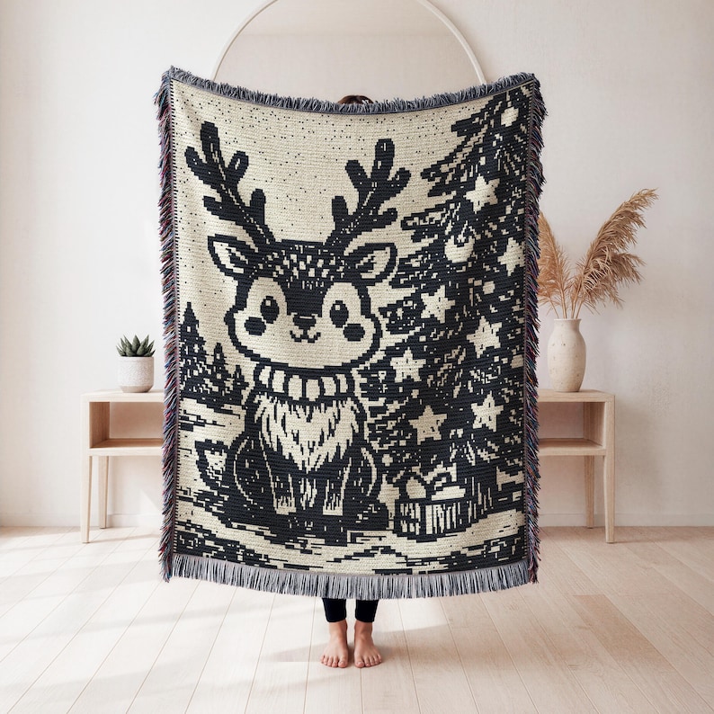 May include: A woven throw blanket featuring a black and cream graphic design of a cartoon reindeer with large antlers, standing next to a decorated Christmas tree with star ornaments and a gift box, finished with a fringed edge on all four sides.