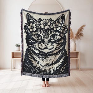 May include: A woven throw blanket featuring a large, high-contrast black and white graphic of a cat wearing a floral crown. The blanket has a textured, knit appearance with a decorative fringe along the top and bottom edges, showcasing a detailed feline face.