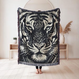May include: A rectangular woven throw blanket featuring a high-contrast black and cream tiger face graphic. The design displays intricate striped patterns and detailed facial features. The edges of the blanket are finished with a fringed border.