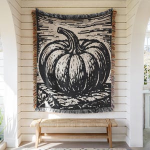 May include: A woven throw blanket featuring a large, black and cream-colored pumpkin graphic centered on the fabric. The rectangular blanket has a fringed edge with multi-colored threads along the left and right sides, displaying a textured pattern.