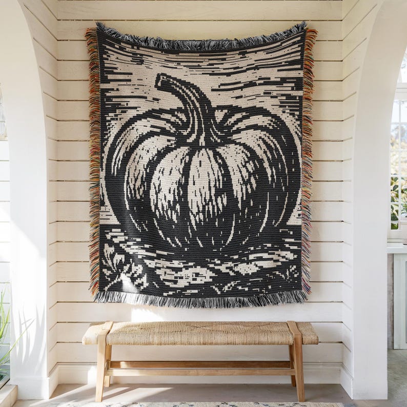 May include: A woven throw blanket featuring a large, black and cream-colored pumpkin graphic centered on the fabric. The rectangular blanket has a fringed edge with multi-colored threads along the left and right sides, displaying a textured pattern.