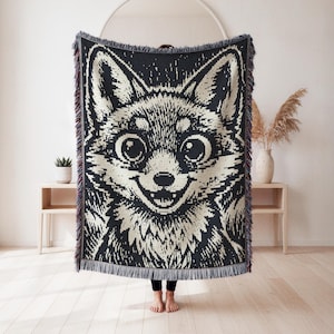 Puede incluir: Una manta tejida rectangular que presenta una ilustración gráfica de alto contraste en negro y crema de una cara de zorro con ojos grandes y orejas puntiagudas. La manta tiene un patrón de punto texturizado y flecos decorativos.