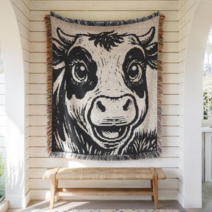 May include: A woven throw blanket featuring a large, high-contrast black and white graphic of a cow's face. The design includes detailed facial features, large eyes, and a wide muzzle. The blanket has a fringed edge with multicolored threads.