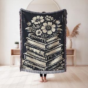 May include: A rectangular woven throw blanket featuring a high-contrast black and cream graphic design. The pattern depicts a stack of five books topped with a bouquet of flowers. The blanket has a decorative fringed edge on all four sides.