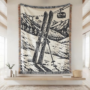 May include: A woven throw blanket featuring a black and cream graphic design of two skis standing upright in snow, a ski pole, and a cable car gondola moving along a line above a mountain landscape, finished with decorative fringed edges.