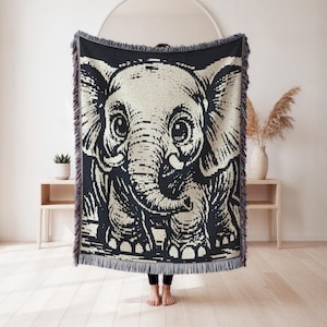 Peut inclure: Un plaid tissé présentant un grand motif détaillé d'un éléphanteau en noir et crème. La couverture a un aspect tricoté texturé avec un bord frangé le long du haut et du bas, mettant en valeur un design à fort contraste visuel.