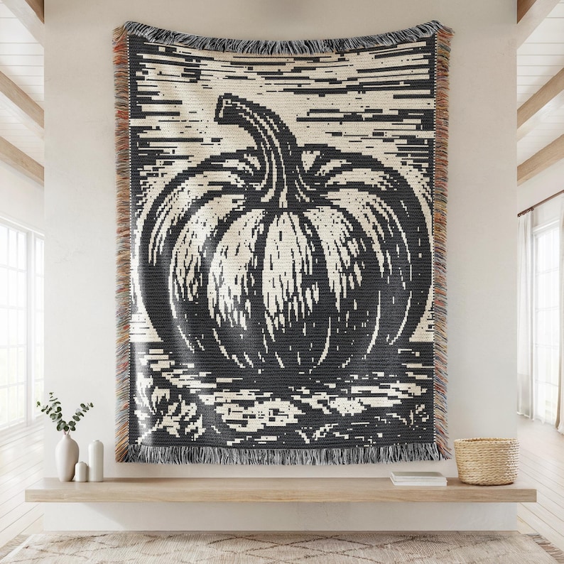 May include: A woven throw blanket featuring a large, black and cream-colored graphic of a pumpkin. The design uses a pixelated, high-contrast style with horizontal lines. The blanket has a fringed edge with multi-colored threads along the sides.
