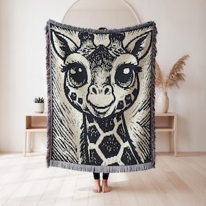 May include: A black and cream-coloured woven throw blanket featuring a large, friendly giraffe face. The giraffe has big eyes and a smiling expression. The blanket has a fringed edge and is held up in front of a person.