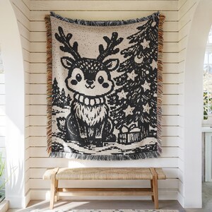 May include: A woven throw blanket featuring a black and cream graphic of a reindeer with antlers standing next to a decorated Christmas tree with star ornaments and a gift box, finished with multicolored fringed edges on the sides.