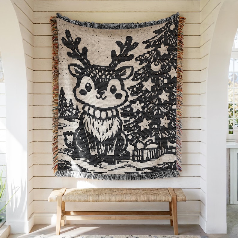 May include: A woven throw blanket featuring a black and cream graphic of a reindeer with antlers standing next to a decorated Christmas tree with star ornaments and a gift box, finished with multicolored fringed edges on the sides.