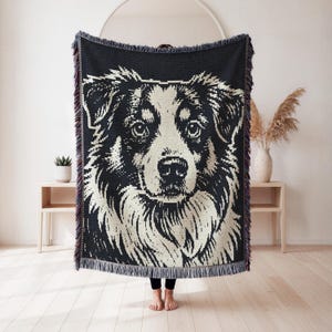 May include: A rectangular woven throw blanket featuring a high-contrast black and cream graphic portrait of an Australian Shepherd dog. The blanket has a textured knit construction and is finished with decorative fringe along the top and bottom edges.