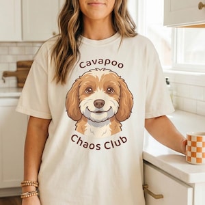 May include: Cream-colored t-shirt featuring a cartoon Cavapoo dog with the text "Cavapoo Chaos Club." The dog has brown and white fur and a smiling expression. The shirt is paired with blue jeans.