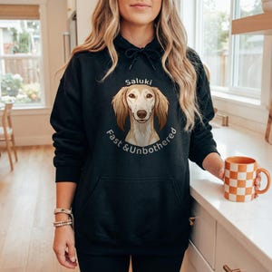 May include: Black hoodie featuring a Saluki dog illustration with the text "Saluki Fast & Unbothered." The hoodie has a front pocket and a drawstring hood. An orange and white checkered mug is held in the hand.