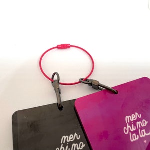 Pode incluir: Dois chaveiros retangulares pretos e roxos com o texto "mer chi mo la la" no chaveiro roxo e "mer chi mo" no chaveiro preto. Os chaveiros est&atilde;o conectados por um cord&atilde;o vermelho.