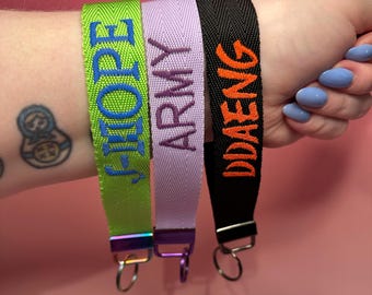 May include: Three colorful keychains with different embroidered text: "J-HOPE", "ARMY", and "JDAENG". The keychains are made of woven fabric and have metal rings.
