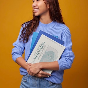 Pode incluir: Uma pessoa jovem vestindo um suéter azul e jeans está segurando uma pilha de livros. O livro de cima tem o título "The Art of Drawing Mermaids: Illustrated Alphabet Guide".