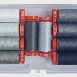 May include: A clear plastic case containing spools of Aurifil cotton thread in various shades of gray, white, and black. The spools have red caps and are labeled with "100% CO" and "MADE IN ITALY". The thread is labeled as No. 50.