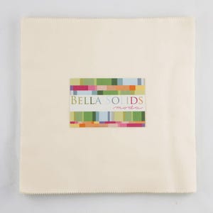 May include: A square, cream-colored fabric piece with a stitched edge. A rectangular label in the center displays the text "BELLA SOLIDS moda" in a colorful, geometric design. The fabric appears to be a quilting material.