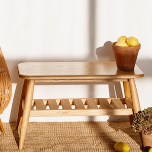 Puede incluir: Un banco de madera clara con un estante de listones y patas anguladas. Una maceta de terracota llena de limones amarillos está sobre el banco. El banco está sobre una alfombra de yute tejida.