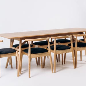 May include: A light wood dining table with six matching chairs. The chairs have black cushioned seats and curved backs. The table and chairs are set against a white background, showcasing a minimalist design.