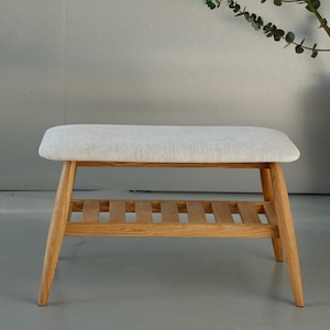 Puede incluir: Un banco tapizado de color claro con estructura de madera. El banco tiene un asiento rectangular y un estante de listones. Las patas están en ángulo y la madera tiene un acabado natural. El banco es de color neutro.