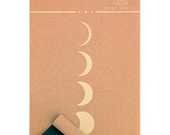 May include: A rectangular yoga mat with a light brown cork surface. The mat features a series of crescent moon phases in a cream colour. The words "PURE SHANTI" are printed at the top. The bottom corner is rolled up, revealing a dark green underside.