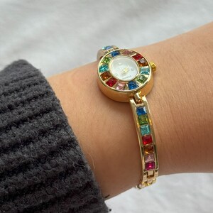 May include: Gold-toned quartz watch with a white face and colorful gemstone accents. The watch features a round face surrounded by a rainbow of gemstones and a matching gemstone bracelet. The watch is worn on a wrist.