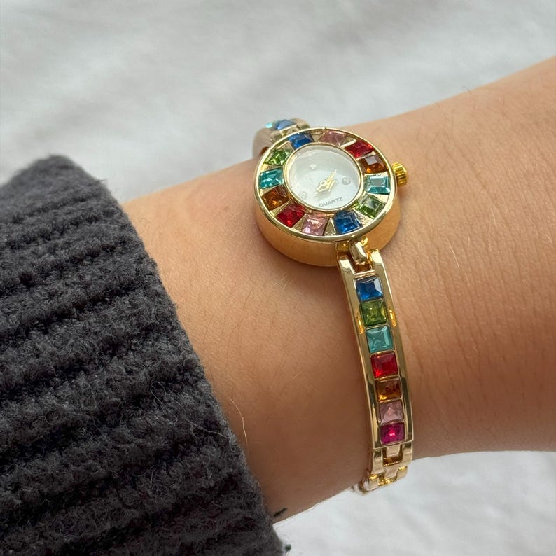 May include: Gold-toned quartz watch with a white face and colorful gemstone accents. The watch features a round face surrounded by a rainbow of gemstones and a matching gemstone bracelet. The watch is worn on a wrist.