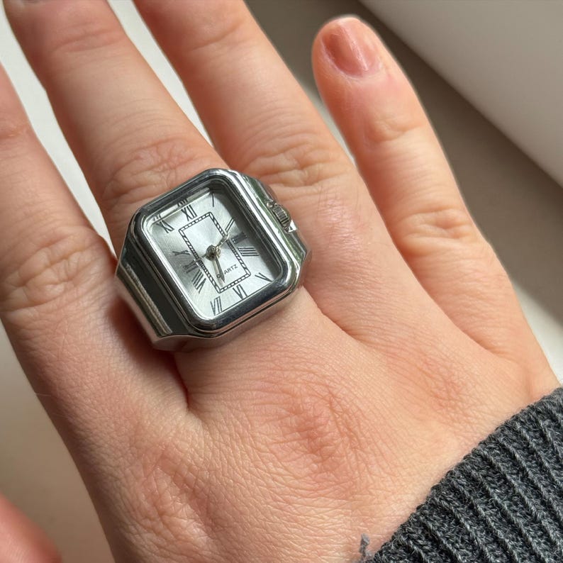 K&ouml;nnte beinhalten: Eine silberfarbene Ringuhr mit quadratischem Zifferblatt und r&ouml;mischen Ziffern. Das Zifferblatt ist hell und die Zeiger sind sichtbar. Der Ring wird am Finger getragen.