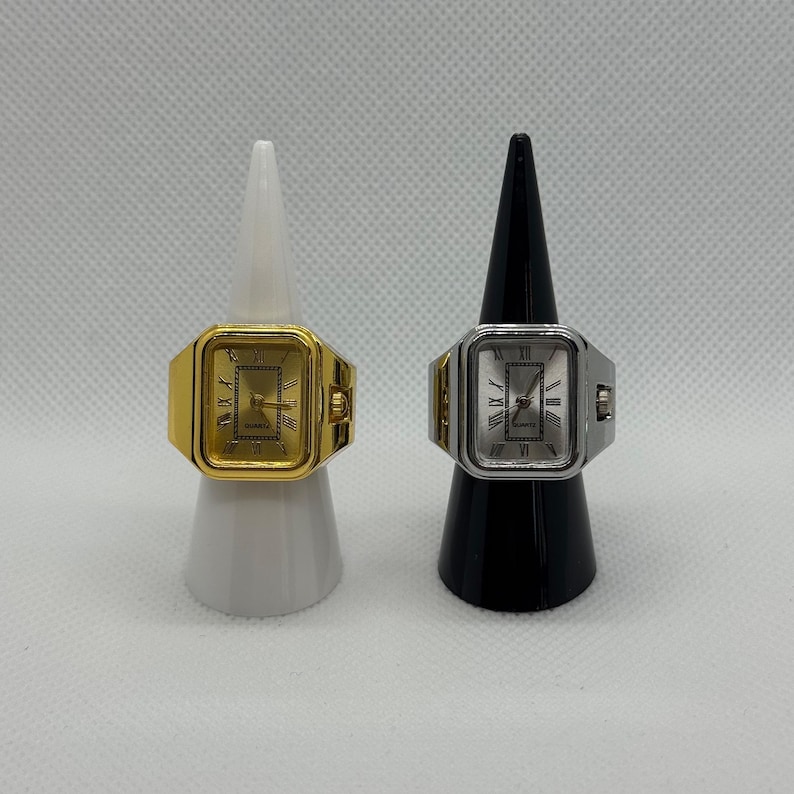 May include: Two ring watches, one gold-toned and one silver-toned, displayed on white and black cone-shaped stands. The watches have rectangular faces with Roman numeral hour markers and hands. The gold watch has a gold face, while the silver watch has a silver face.