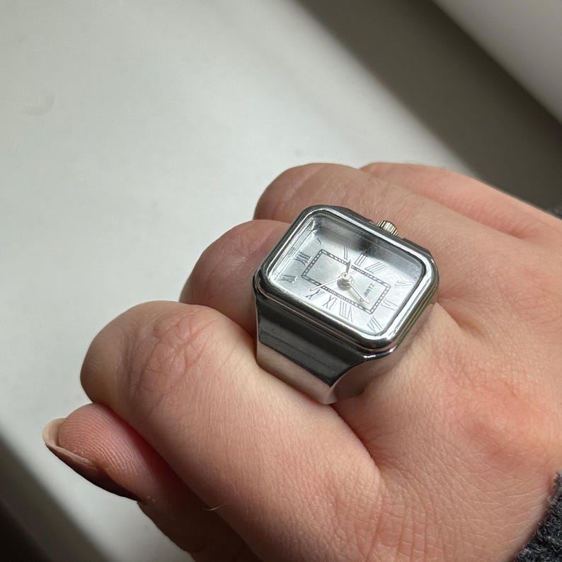 K&ouml;nnte beinhalten: Ein silberfarbener Ring mit rechteckigem Zifferblatt. Das Zifferblatt der Uhr hat r&ouml;mische Ziffern und silberne Zeiger. Der Ring wird an einem Finger getragen.
