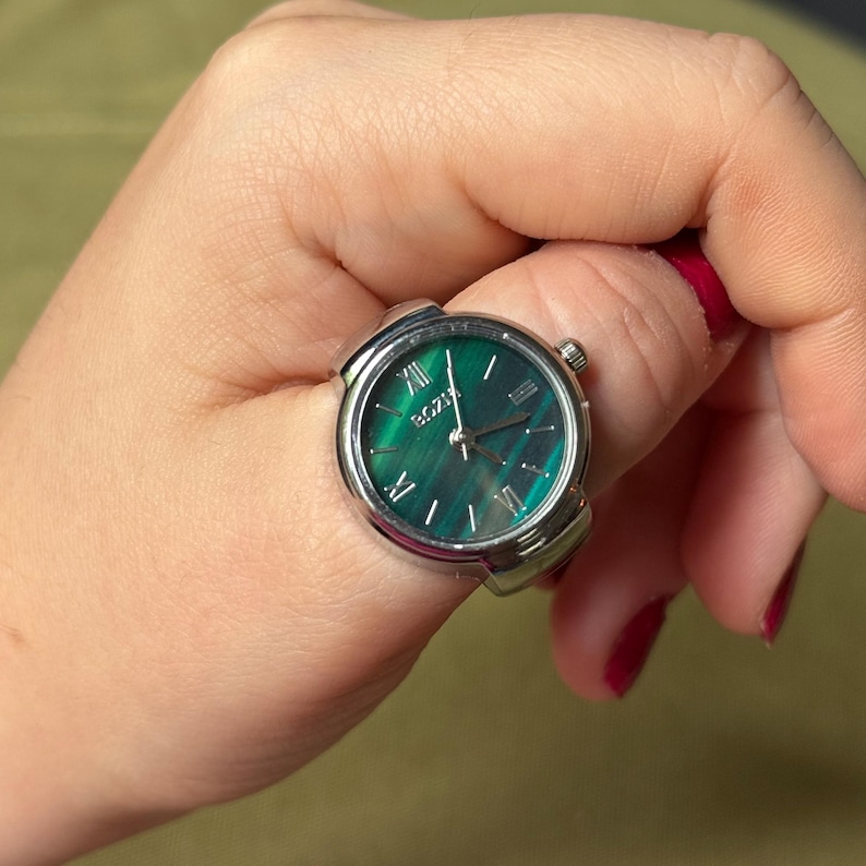 May include: A silver ring watch with a green face. The watch face has Roman numerals and the brand name "BOZE". The watch is being worn on a finger.