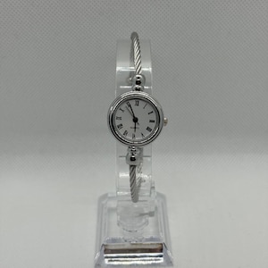 Peut inclure: Une montre à quartz argentée avec un cadran rond et des chiffres romains. La montre a un bracelet argenté avec un design torsadé. La montre est présentée sur un présentoir en plastique transparent.