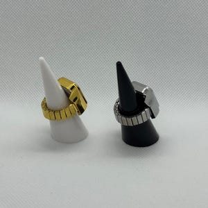 May include: Two rings on display. One is gold-toned, the other is silver-toned. Both have a rectangular face and a band made of small rectangular segments. The gold ring is on a white cone, and the silver ring is on a black cone.