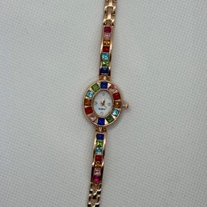 May include: A rose gold quartz watch with a white face and colorful square gemstones. The watch band and the bezel around the watch face are adorned with a variety of colorful gemstones. The watch face has the word "QUARTZ" printed on it.