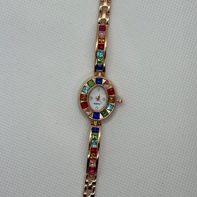 May include: A rose gold quartz watch with a white face and colorful square gemstones. The watch band and the bezel around the watch face are adorned with a variety of colorful gemstones. The watch face has the word "QUARTZ" printed on it.