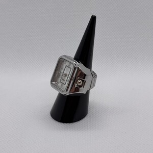 K&ouml;nnte beinhalten: Eine silberfarbene Ringuhr mit rechteckigem Zifferblatt und r&ouml;mischen Ziffern. Die Uhr wird auf einem schwarzen Kegel pr&auml;sentiert. Das Zifferblatt ist wei&szlig; mit silbernen Zeigern und dem Wort "QUARTZ" unten.