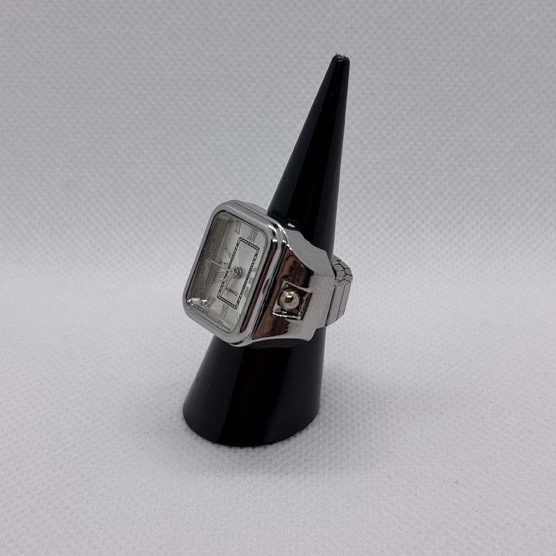 K&ouml;nnte beinhalten: Eine silberfarbene Ringuhr mit rechteckigem Zifferblatt und r&ouml;mischen Ziffern. Die Uhr wird auf einem schwarzen Kegel pr&auml;sentiert. Das Zifferblatt ist wei&szlig; mit silbernen Zeigern und dem Wort "QUARTZ" unten.