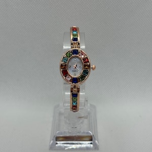 May include: A rose gold quartz watch with an oval face and a band adorned with colorful gemstones. The watch face is white with black hands and the word "QUARTZ" printed at the bottom. The watch is displayed on a clear stand.