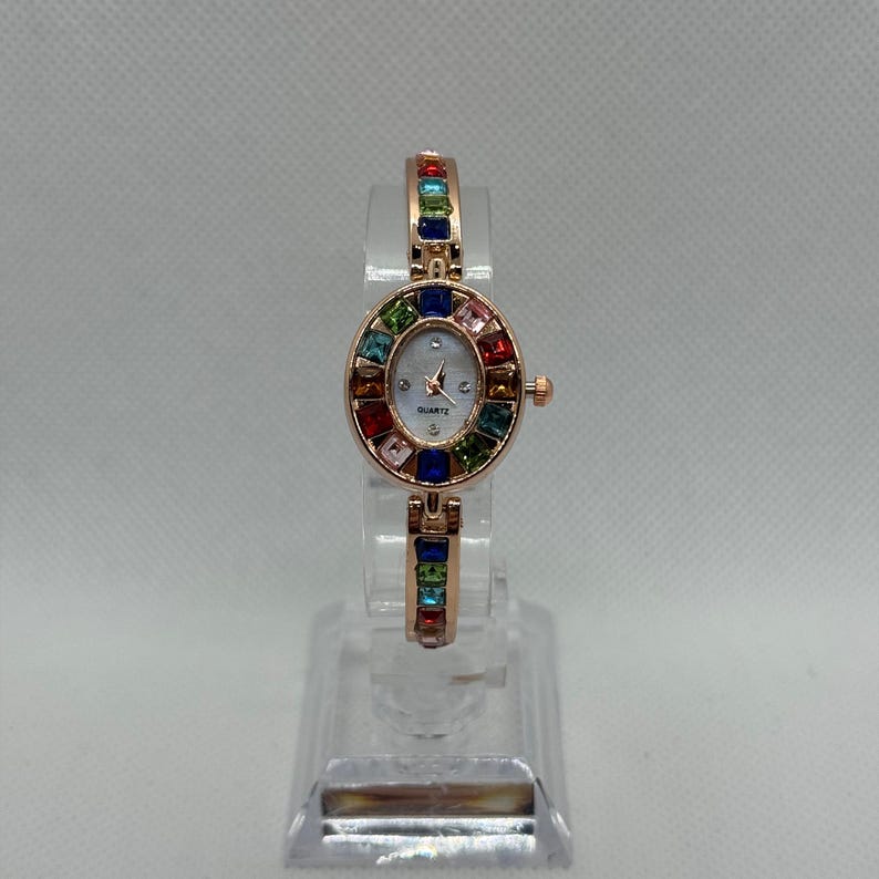 May include: A rose gold quartz watch with an oval face and a band adorned with colorful gemstones. The watch face is white with black hands and the word "QUARTZ" printed at the bottom. The watch is displayed on a clear stand.