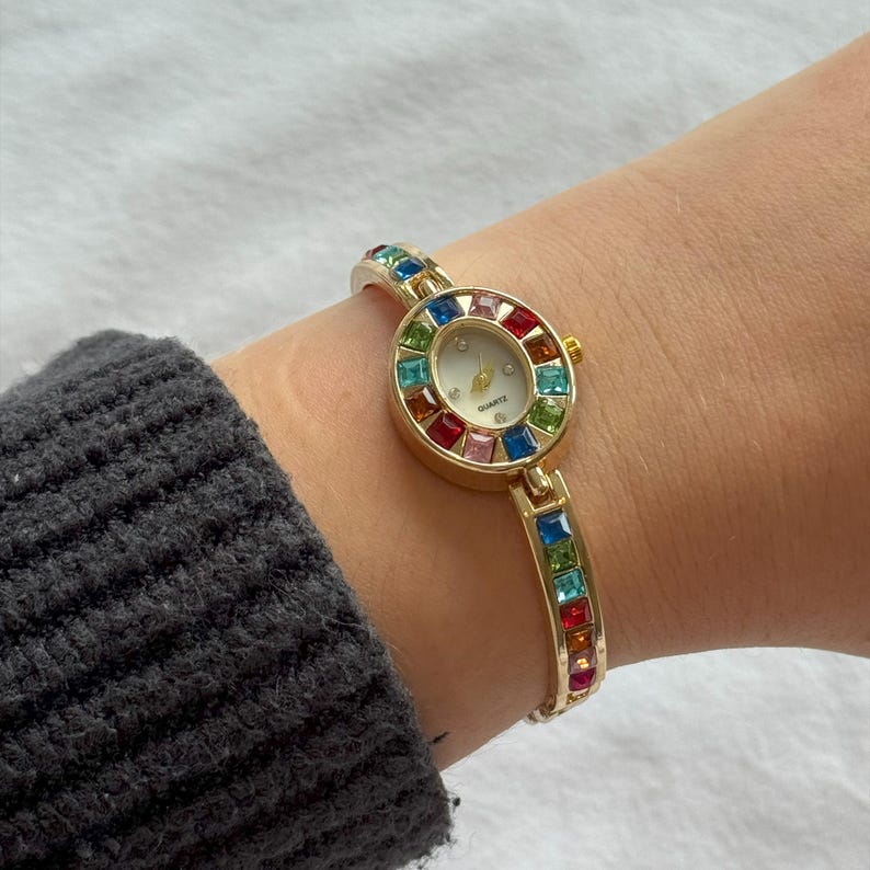 May include: Gold-toned quartz watch with a round face and a band adorned with colorful gemstones. The watch face is white with gold hands and the word "Quartz" printed on it. The band is a gold color.