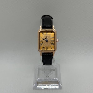 May include: A gold-toned rectangular wristwatch with a black leather band. The watch face features Roman numerals and a textured center. The watch is displayed on a clear plastic stand.