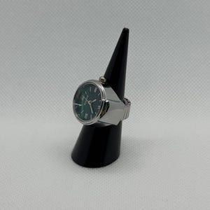 May include: A silver ring watch with a dark green face and silver hands. The watch has a round face with Roman numerals and is displayed on a black cone-shaped stand. The band is expandable.